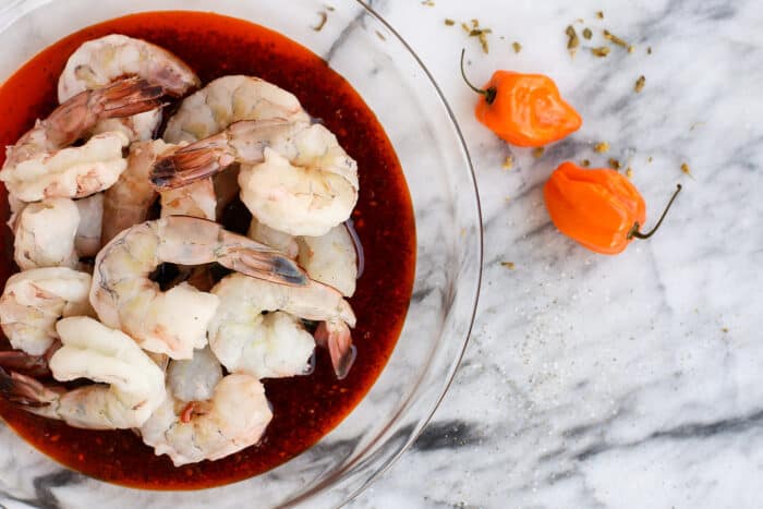 Overhead view of raw shrimp in a bowl of marinade next to two habanero peppers.