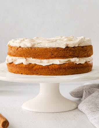 a pumpkin flavored double layer cake with creamy cinnamon whipped topping on a white cake stand