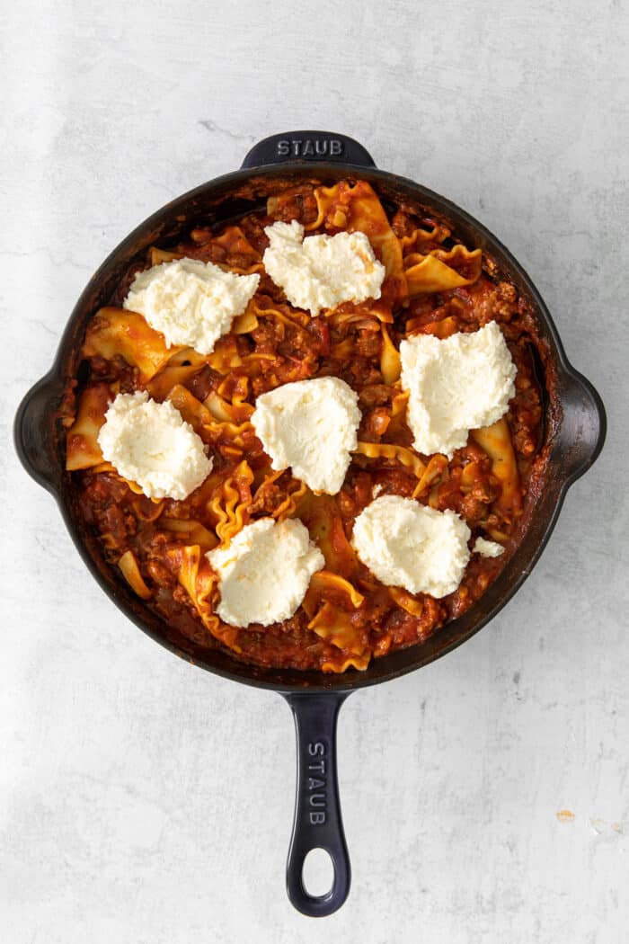 Dollops of ricotta cheese over lasagna noodles and sauce in a skillet