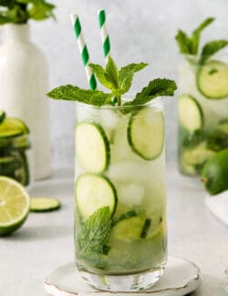 A tall glass of cucumber mojito garnished with cucumber and mint and finished with two green straws.
