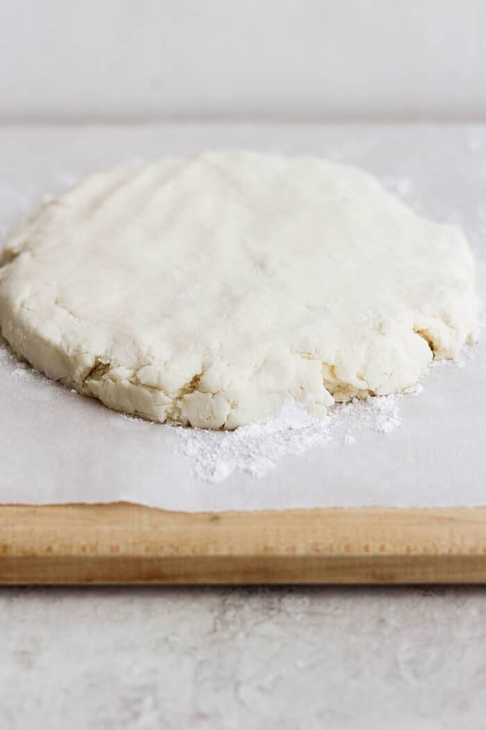A round of cream biscuit dough rests on a white piece of parchment paper on top of a wooden cutting board.