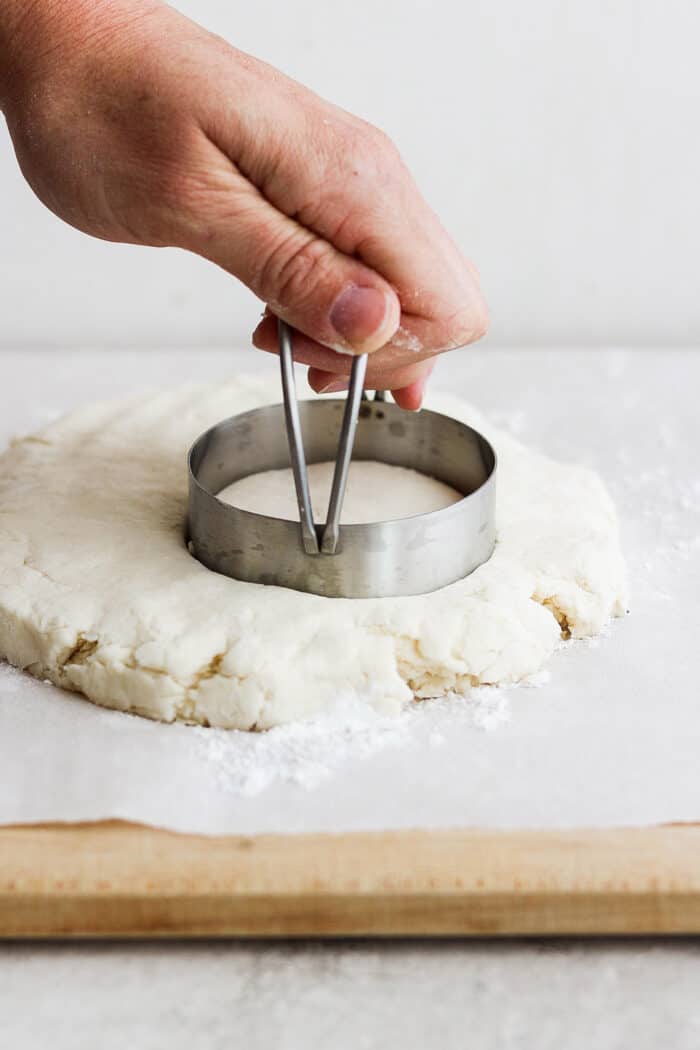 A hand uses a metal biscuit cutter to cut out a cream biscuit from a piece of dough.