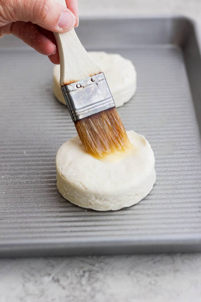 A hand uses a pastry brush to brush the top of a cream biscuit before it's baked.