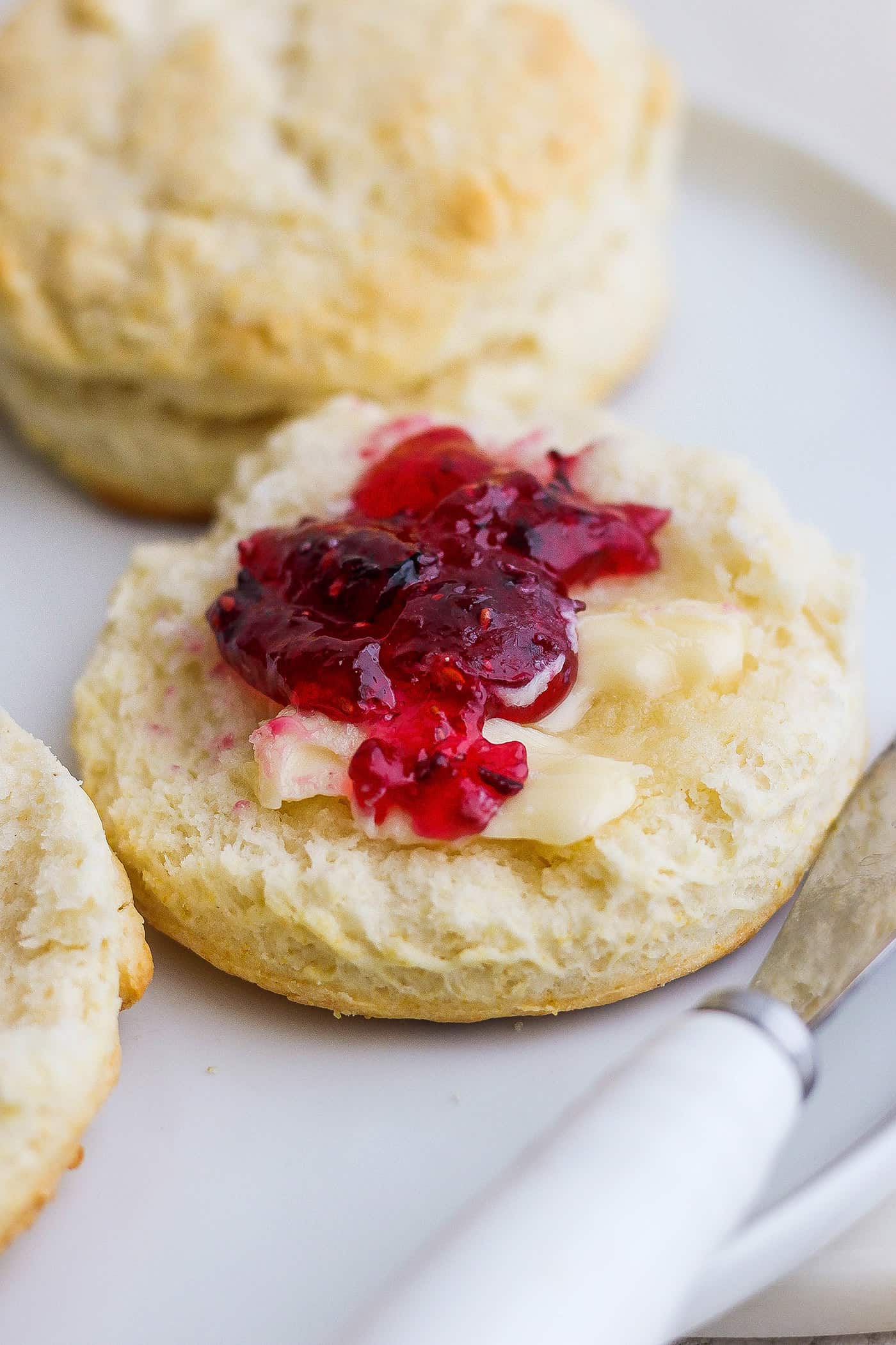 A cut cream biscuit is shown spread with butter and jam with more biscuits in the background.