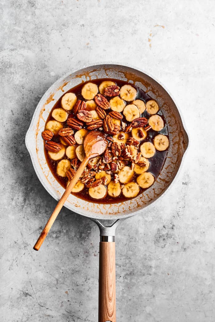 A wooden spoon stirs a skillet of cooking banana slices for bananas foster.