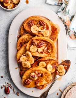 An oval serving dish holds slices of bananas foster French toast topped with bananas.