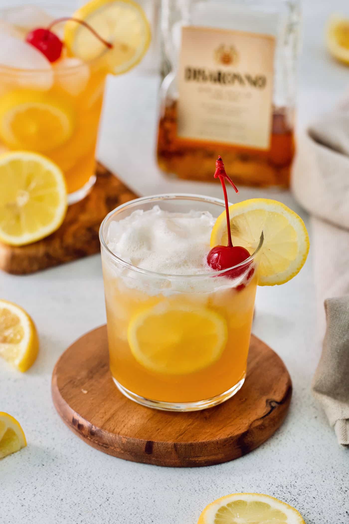 Glasses of amaretto sour are garnished with lemon slices and cherries, with a bottle of amaretto shown in the background.