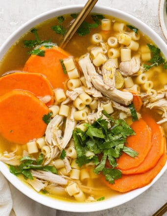A hearty bowl of ginger chicken soup showing shredded chicken, sliced sweet potatoes, and pasta.