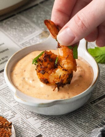 A hand dips a piece of blackened shrimp into a bowl of shrimp dipping sauce.