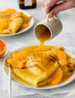 A hand pours a pitcher of orange sauce over a plate of crepes Suzette.