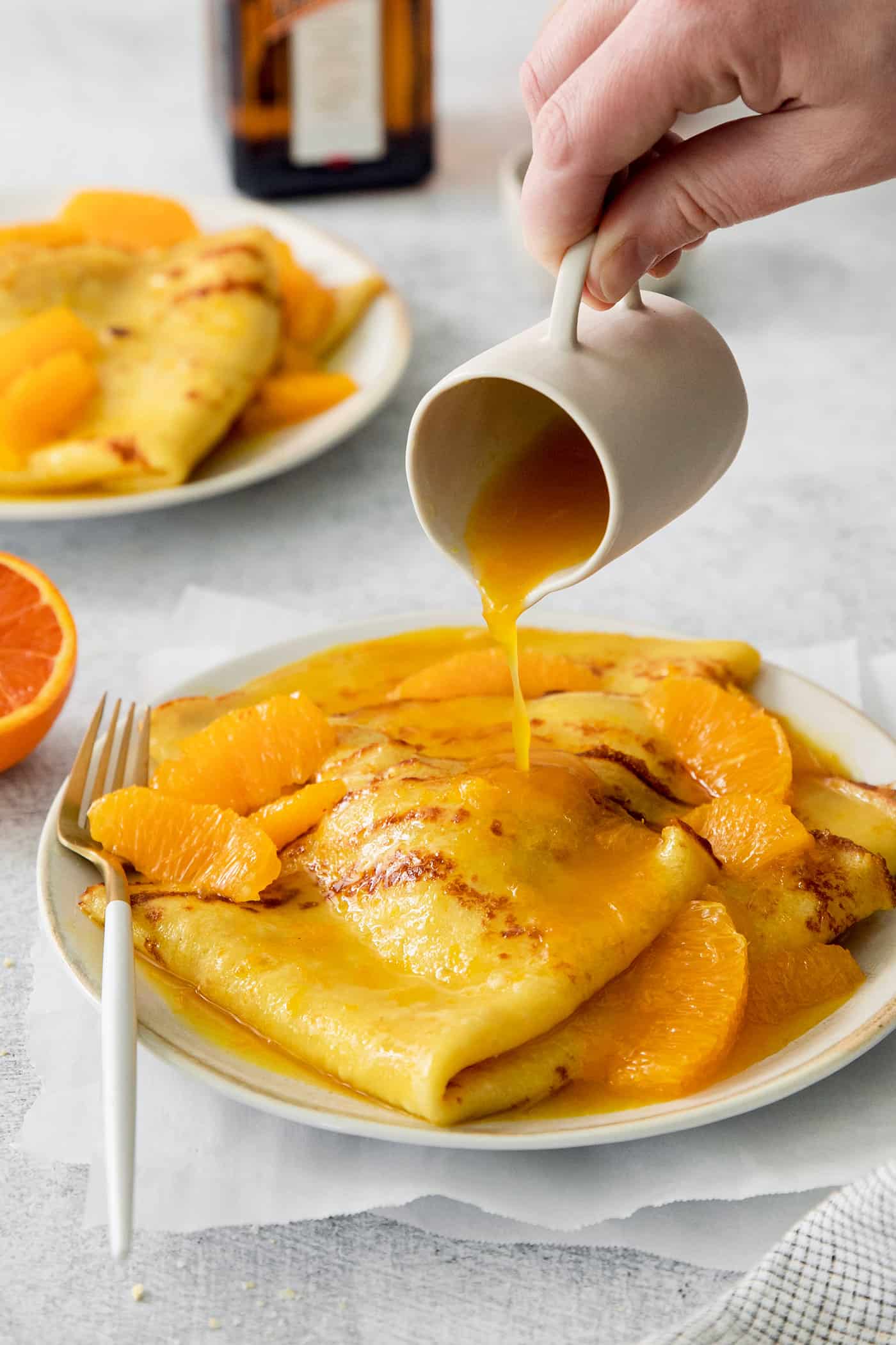 A hand pours a pitcher of orange sauce over a plate of crepes Suzette.
