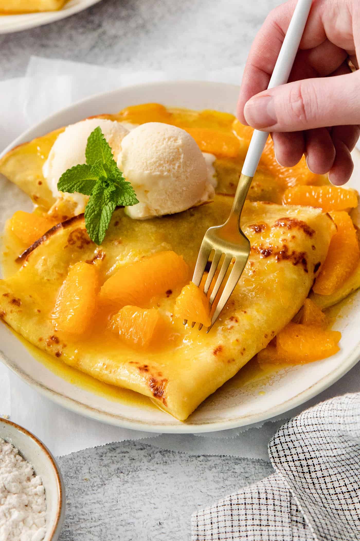 A scoop of vanilla ice cream tops crepes Suzette as a fork cuts into them.
