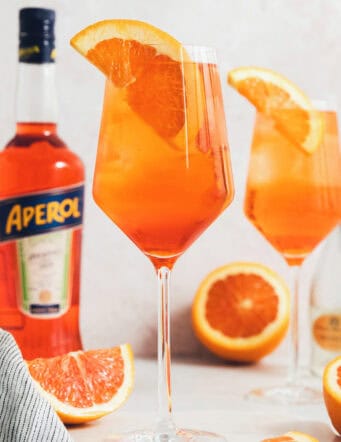 Glasses of Aperol spritz are surrounded with orange wedges next to a bottle of Aperol.