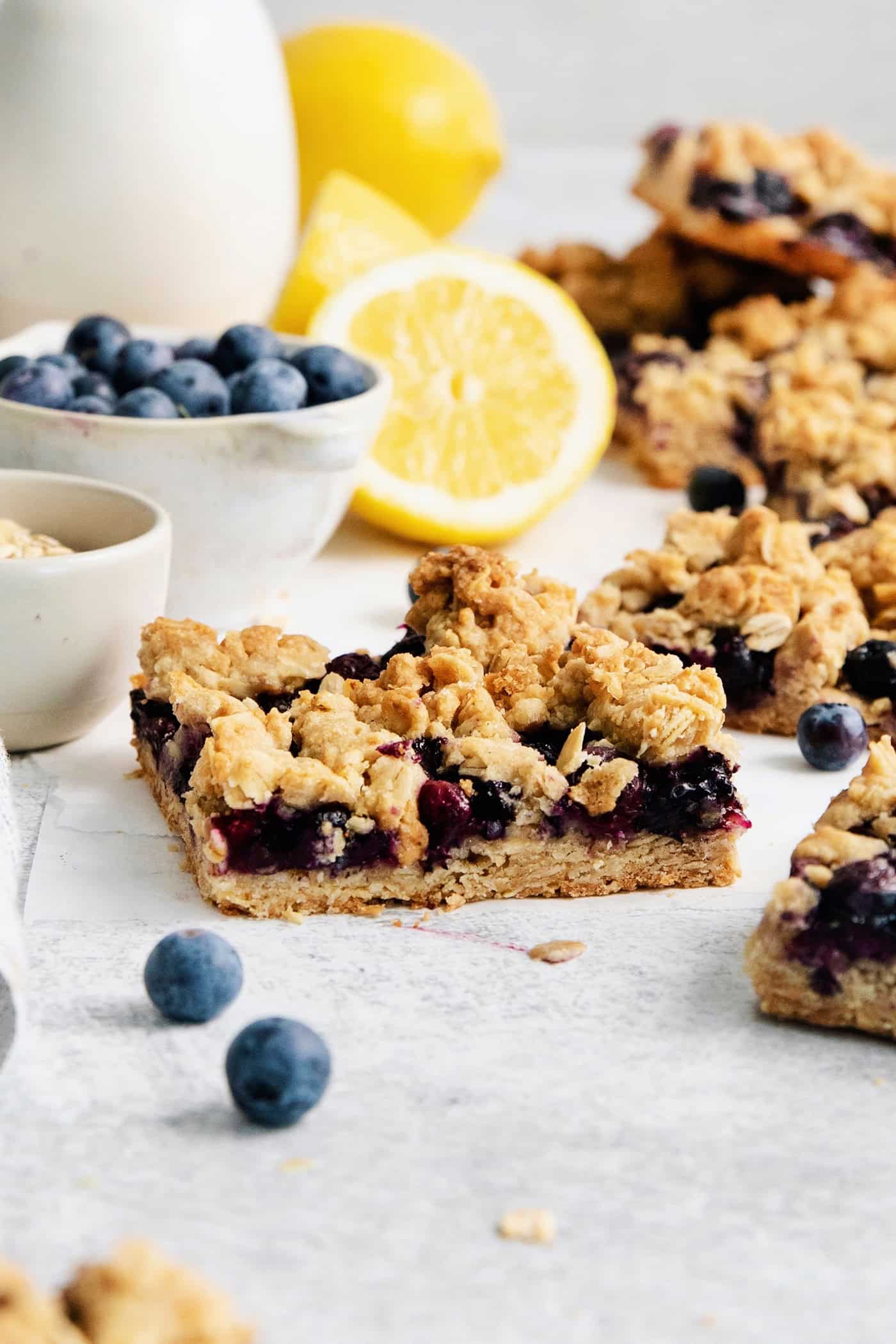 Blueberry crumble bars surrounded by fresh lemons and bowls of blueberries.