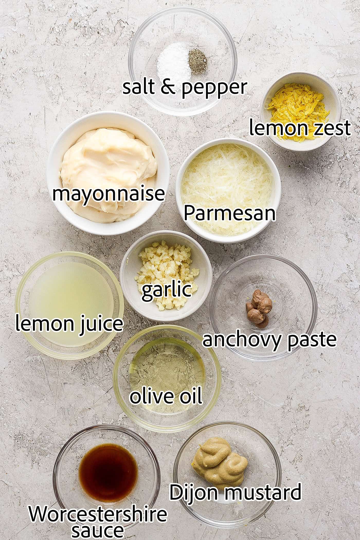 Ingredients for caesar dressing are shown portioned out and labelled: lemon zest and juice, mayonnaise, parmesan cheese, anchovy paste, salt and pepper, Worcestershire sauce, mustard, olive oil.