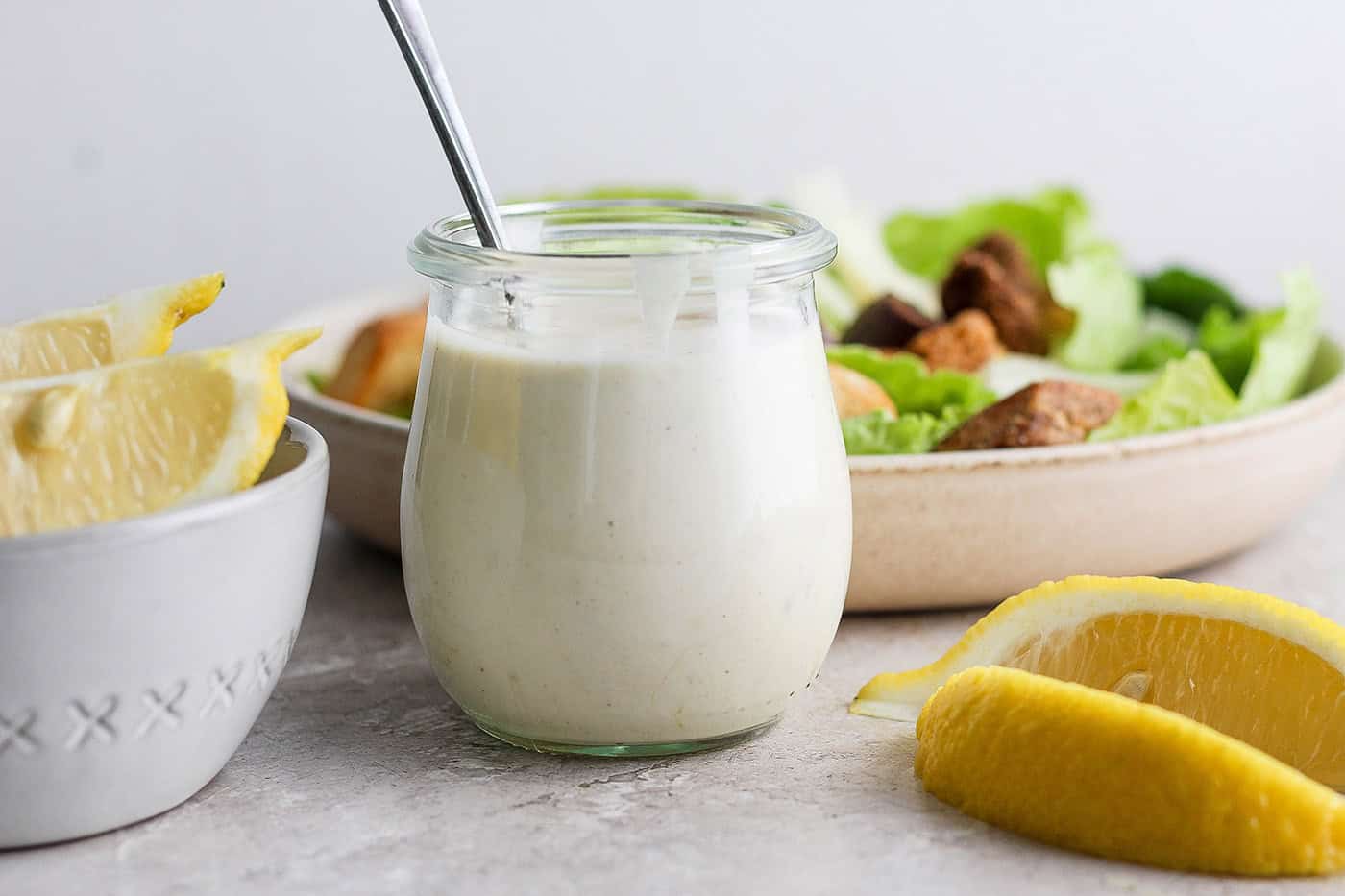 A glass jar of caesar dressing with a spoon in it surrounded by salad.