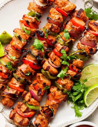 Four grilled charmoula pork kabobs on a plate with lime wedges.