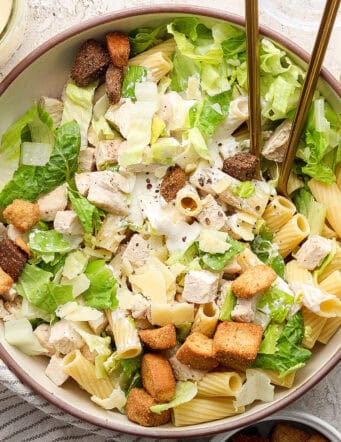 A big bowl of Caesar chicken pasta salad with serving utensils.