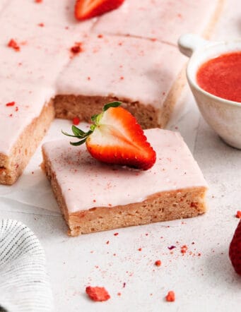 Pink icing-topped strawberry brownies are cut into squares, with strawberries scattered around them.